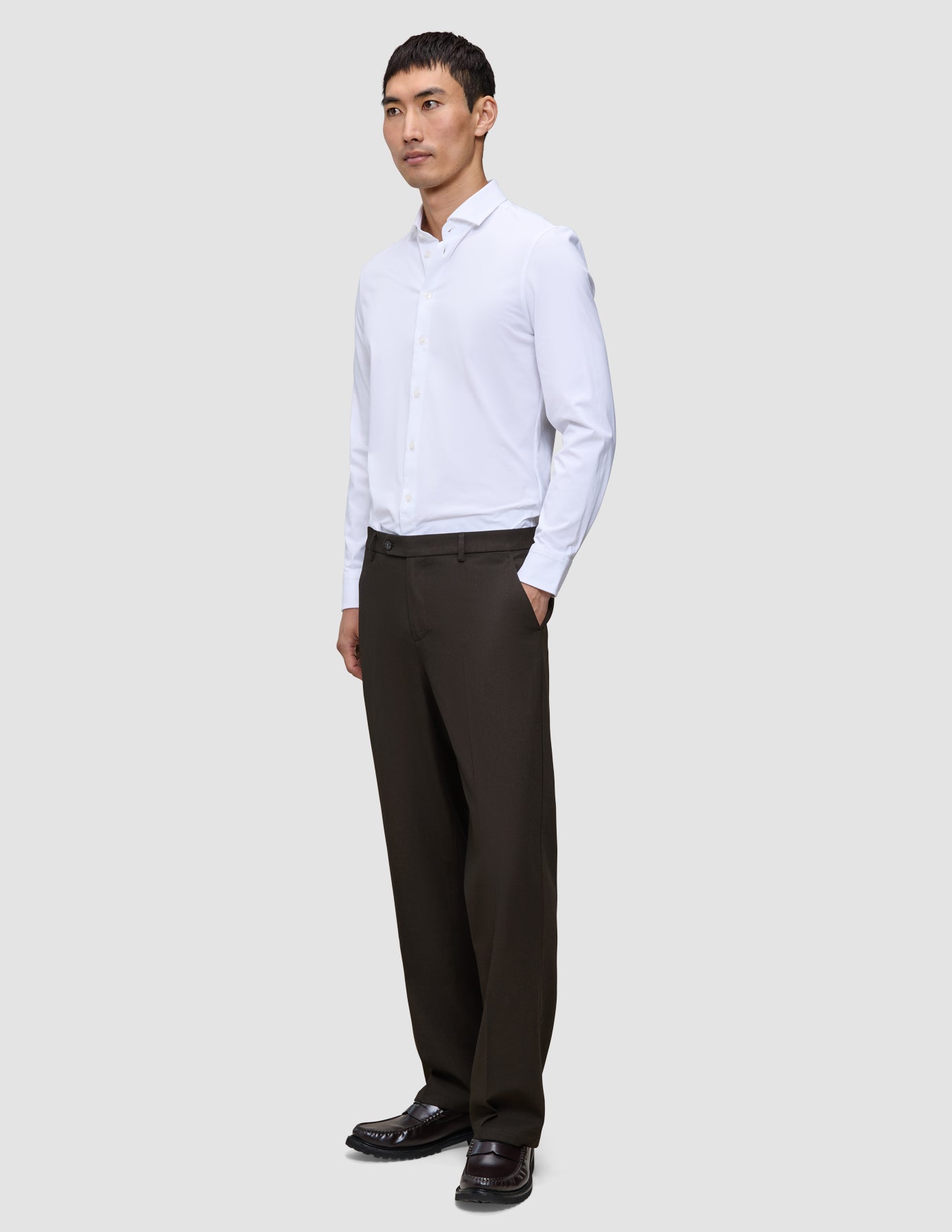 Ein Mann in einem weißen Hemd und einer braunen, locker sitzenden Motion Suit-Hose Relaxed Cognac steht da, eine Hand in der Hosentasche.